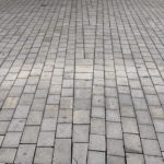 Graue rechteckige Pflastersteine, die zu einem NATURSTEIN-PFLASTER-SYSTEM gehören, sind in einem weitgehend einheitlichen Muster auf dem Gehweg verlegt. Einige Steine sind uneben oder leicht erhöht. Im Hintergrund sind zwei schwarze Mülltonnen und grünes Laub zu sehen.